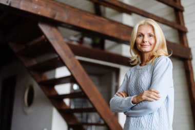 Orta yaşlı gülümseyen sarışın kadın modern odada duruyor.