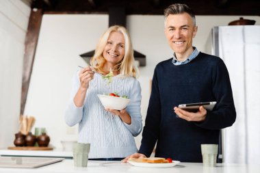 Mutlu orta yaşlı çift evde tabletle web sörfü yapıyor, kahvaltı yapıyor, masada ayakta duruyor.