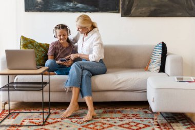 Beyaz anne ve kız evdeki kanepede otururken cep telefonu ve kulaklık kullanıyor.