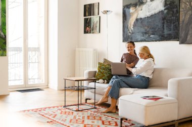 Evdeki koltukta otururken gülümseyen ve cep telefonu kullanan beyaz bir anne ve kız.