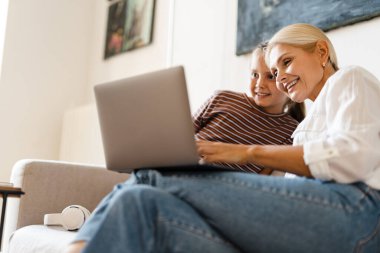 Evdeki koltukta otururken gülümseyen ve cep telefonu kullanan beyaz bir anne ve kız.