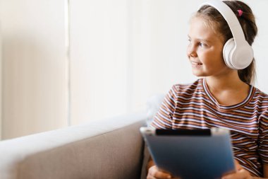 Kulaklıklı beyaz kız evde koltukta otururken tablet bilgisayar kullanıyor.