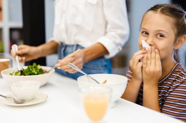 Beyaz anne ve kız mutfakta yemek yerken gülümsüyor.