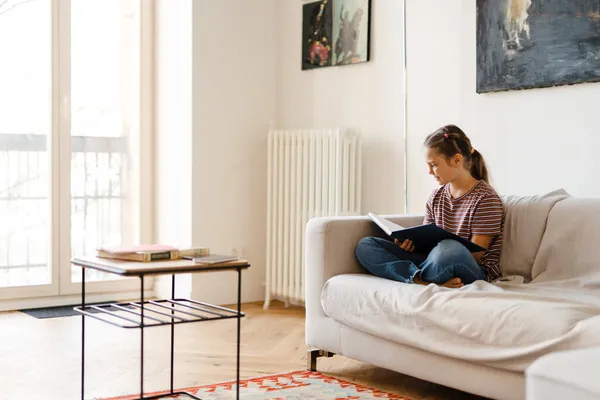 Gadis Pra Remaja Tersenyum Dan Membaca Buku Sambil Duduk Sofa — Stok Foto