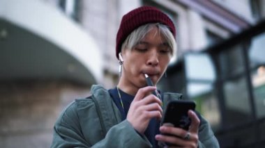 Yakışıklı Asyalı adam dışarıda sigara içiyor ve telefonla mesajlaşıyor.