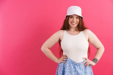 Dövmeli genç kızıl kadın gülümsüyor ve pembe arka planda izole edilmiş kameraya bakıyor.