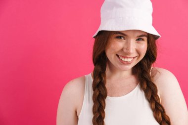 Saçları örgülü, gülümseyen ve pembe arka planda izole edilmiş kameraya bakan kızıl saçlı genç bir kadın.