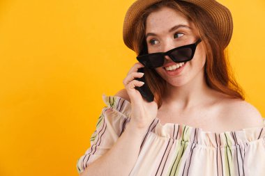 Sarı arka planda izole edilmiş, gülümseyen ve cep telefonuyla konuşan şapkalı genç kızıl kadın.