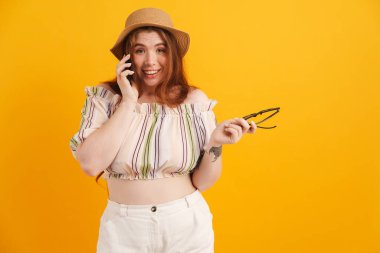 Sarı arka planda izole edilmiş, gülümseyen ve cep telefonuyla konuşan şapkalı genç kızıl kadın.