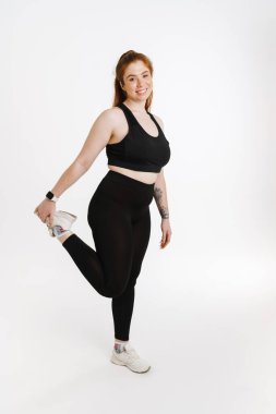 Gülümseyen genç ve beden ölçülerinde spor kıyafetli bir kadın beyaz arka planda durup esneme egzersizleri yapıyor.