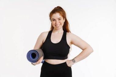 Beyaz arka planda izole edilmiş spor minderiyle poz verirken gülümseyen kızıl saçlı genç kadın.