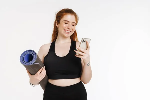Junge Ingwerfrau Benutzt Handy Während Sie Mit Fitnessmatte Isoliert Vor — Stockfoto