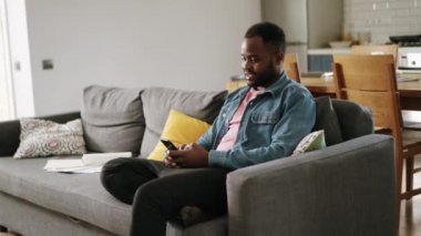 Afrikalı mutlu adam evde, kanepede telefonla mesajlaşıyor.