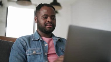 Evdeki kanepede dizüstü bilgisayarla yazan neşeli Afrikalı adam.