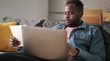 Evdeki kanepede not defterinden dizüstü bilgisayara bir şeyler yazan Afrikalı bir adam.