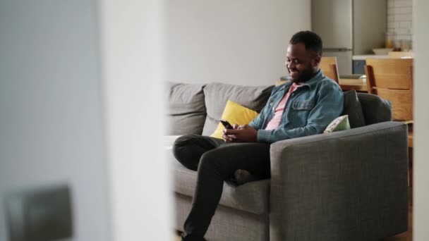 アフリカの笑い男はソファの上で自宅で電話でテキストメッセージ — ストック動画