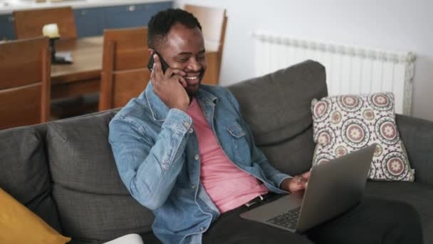 陽気なアフリカの男はラップトップを見て ソファの上に自宅で電話で話す — ストック動画