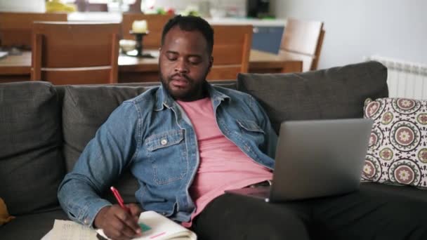 ソファの上の自宅でノートパソコンから何かを書くアフリカの男 — ストック動画