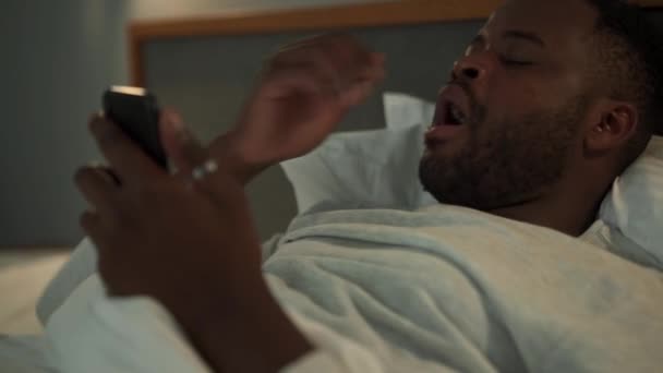 疲れてアフリカ人の男があくびをし ベッドの上で電話を見て — ストック動画
