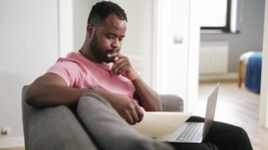 Afrikalı dalgın adam evdeki kanepede gazeteye bakıyor.