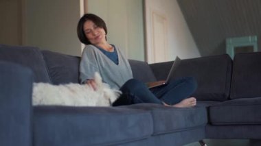 Bilgisayarda çalışan ve köpeğini okşayan gülümseyen olgun bir kadın.