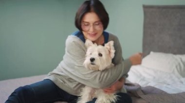 Düşünceli olgun bir kadın köpeğini yatakta kucaklıyor.