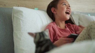 Küçük Asyalı kız telefona bakıyor ve kedisini yatakta okşuyor.