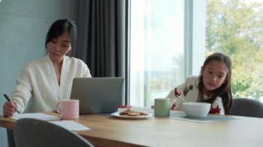 Kızı sabah bir şey isterken Asyalı düşünceli anne dizüstü bilgisayarda çalışıyor.