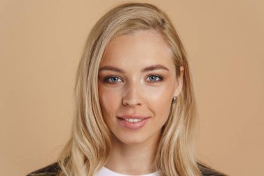 Bej arka planda durup kameraya bakan uzun saçlı, gülümseyen sarışın bir kadının güzel görüntüsü.