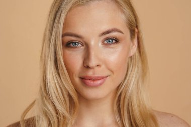 Bej arka planda durup kameraya bakan uzun saçlı, gülümseyen sarışın bir kadının güzel görüntüsü.