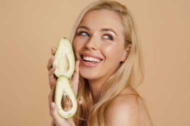 Sırıtan sarışın, üstsüz, uzun saçlı, bej arka planda duran, elinde avokado tutan kadın. 