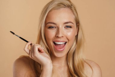 Gülümseyen, beyaz, sarışın, çıplak omuzlu, bej arka planda duran, elinde kaş fırçası olan bir kadın.