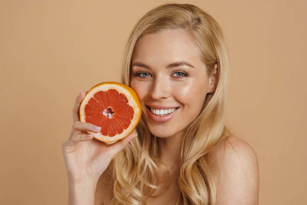 stock image Smiling young blonde topless woman with long hair standing over beige background, holding cut grapefruit