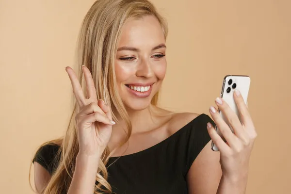 笑着拿着手机站在米黄色背景旁挥手视频通话的年轻白人金发女人 — 图库照片