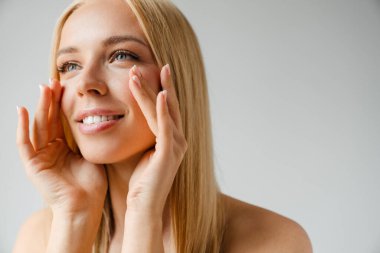 Tişörtsüz sarışın kadın tenine dokunuyor ve beyaz arka planda soyutlanmış bir şekilde yan bakıyor.