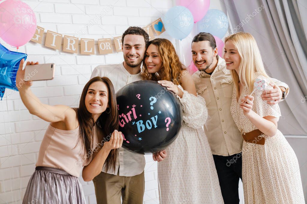 Happy young pregnant woman celebrating baby shower with her friends at home, taking selfie together, holding balloon