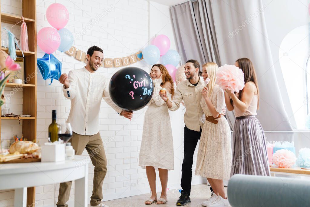 Happy young pregnant woman with friends at a baby shower, poping gender balloon
