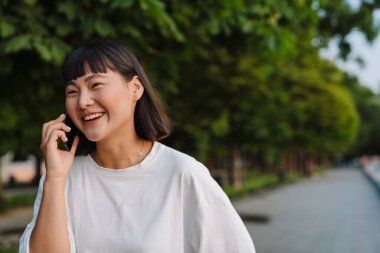 Yeşil bir parkta yürüyen gülümseyen Asyalı genç kadın cep telefonuyla konuşuyor.