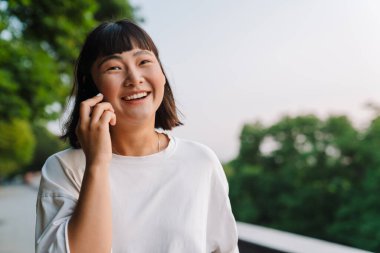 Yeşil bir parkta yürüyen gülümseyen Asyalı genç kadın cep telefonuyla konuşuyor.