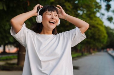 Gülümseyen Asyalı genç bir kadın yeşil bir parkta yürüyor kablosuz kulaklıklarla müzik dinlemekten zevk alıyor.