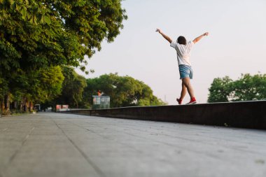 Yeşil bir parkta yürüyen, kulaklıklı Asyalı genç bir kadının arka görüntüsü.