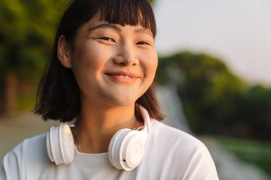 Gülümseyen Asyalı genç bir kadın yeşil bir parkta yürüyor kablosuz kulaklıklarla müzik dinlemekten zevk alıyor.