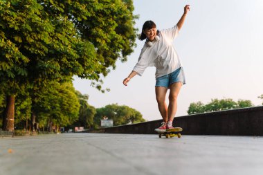 Yeşil parkta kaykay süren mutlu genç Asyalı kadın.