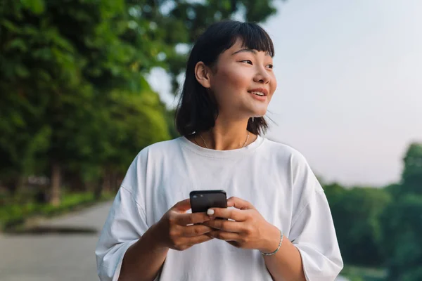 Uśmiechnięta Młoda Azjatka Spacerująca Zielonym Parku Pomocą Wiadomości Przez Telefon — Zdjęcie stockowe