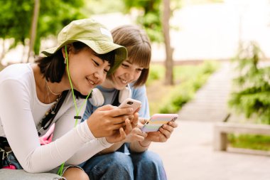 Çok ırklı iki kadın yeşil parkta dinlenirken cep telefonu kullanıyor ve gülümsüyor.