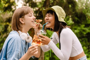 Çok ırklı iki kadın yeşil parkta dinlenirken kokteyl içip pizza yiyor.