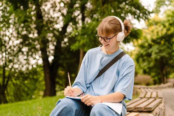 Kulaklıkla Müzik Dinleyen Genç Bir Kadın Parkta Otururken Notlar Yazıyor — Stok fotoğraf