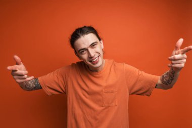 Kırmızı duvarın arkasında durup kamerayı işaret eden uzun kahverengi saçlı, gülümseyen beyaz, sıradan bir adamın portresi.