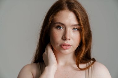 Duvarın arkasında durup kameraya bakan uzun kızıl saçlı, kıvrımlı genç bir kadının portresi.