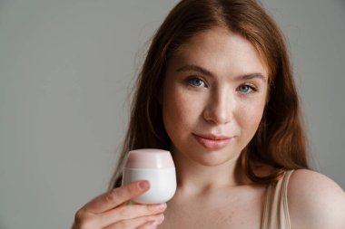 Gri duvarın üzerinde duran krema kavanozunu gösteren gülümseyen genç bir kadının yakın çekimi.